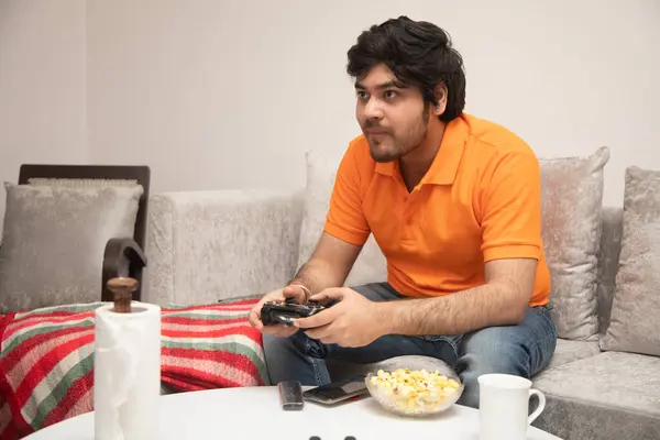 Asyalı Hintli adam oyun oynuyor, canlı yayın yapıyor ve TV seyrediyor. Oturma odasında, gri bir kanepe ve beyaz bir masa üzerinde..