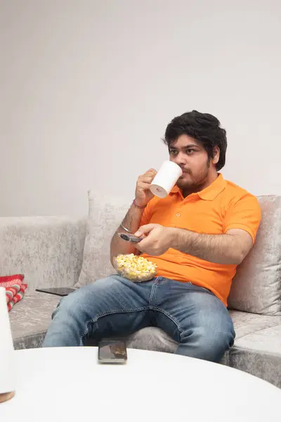 Asyalı Hintli adam oyun oynuyor, canlı yayın yapıyor ve TV seyrediyor. Oturma odasında, gri bir kanepe ve beyaz bir masa üzerinde..
