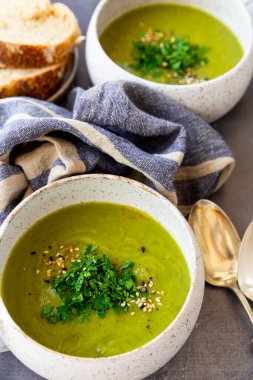 Beyaz seramik malzemeli yeşil çorba, peçeteler, kaşık, gri bir yüzey üzerinde ekmek dilimleri.