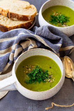 Beyaz seramik malzemeli yeşil çorba, peçeteler, kaşık, gri bir yüzey üzerinde ekmek dilimleri.