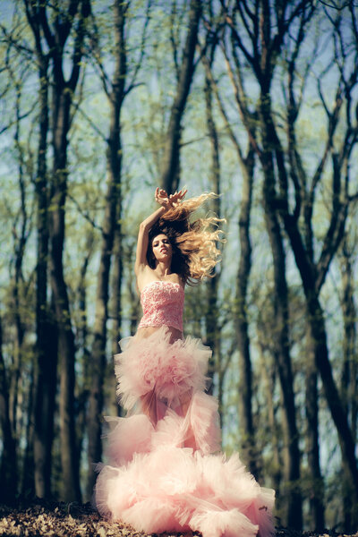 Beautiful young woman in pink glamour dress with long curly hair dancing in sunny forest