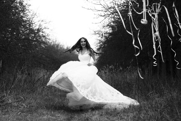 Happy bride with in white wedding dress dancing outdoor near trees with decorative ribbons, black and white