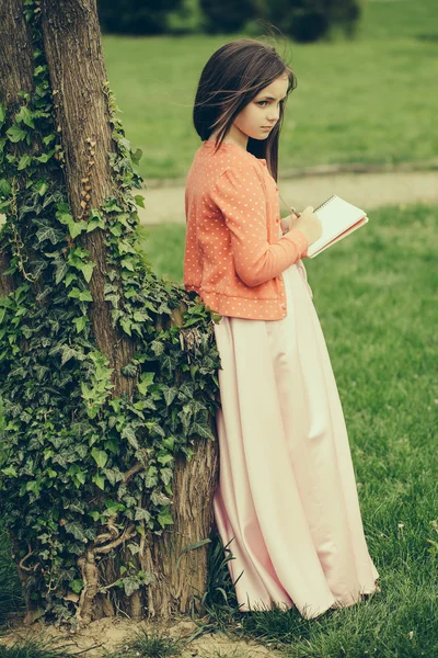 Menina em vestido com caderno e caneta perto da árvore — Fotografia de Stock