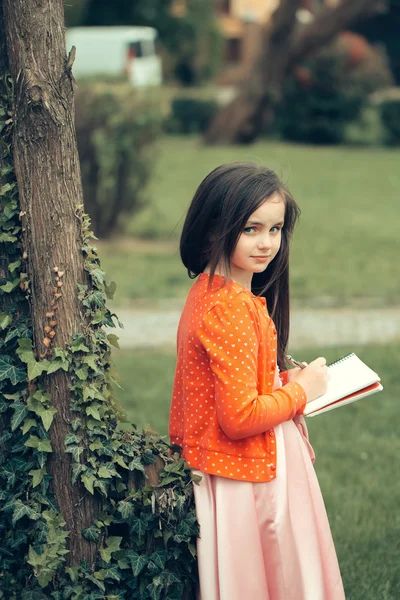 Menina em vestido com caderno e caneta perto da árvore — Fotografia de Stock