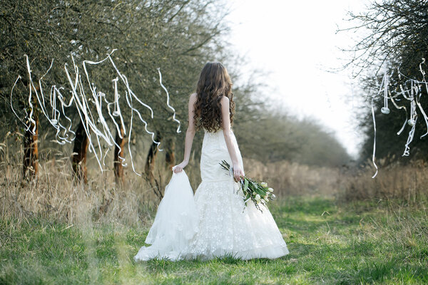 Beautiful young bride in white wedding dress with flowers in field with ribbons on trees outdoor