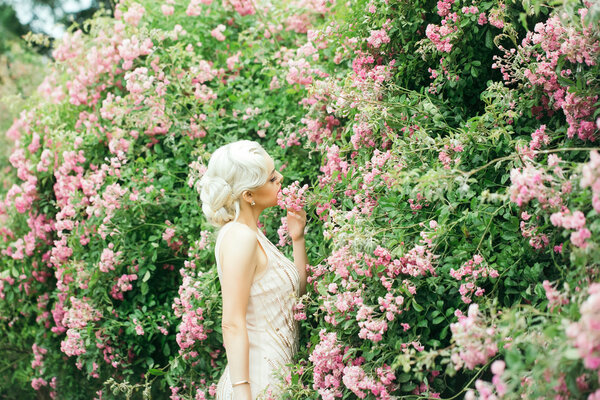 Young girl with blonde hair fashion makeup on pretty face in beautiful cream-colored dress posing near pink rose bush in bloom