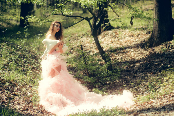 Young woman with beautiful face and long curly hair in glamour pink dress holding cute white small goat in sunny forest