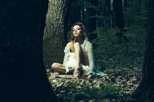 Young woman with beautiful face sitting in soft knitted blue cloth holding cute white small goat in deep forest