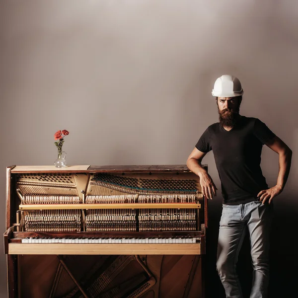 Hombre barbudo en casco cerca de piano de madera — Foto de Stock