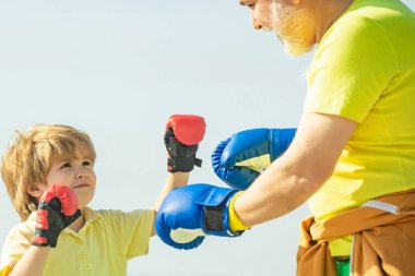 Boks tekmesi pratiği yapan yakışıklı yaşlı adam. Yaşlı adam kum torbasına vuruyor. Büyükbaba ve çocuk dövüşü pozları. Kıdemli antrenör ve boks eldiveni giyen küçük çocuk..