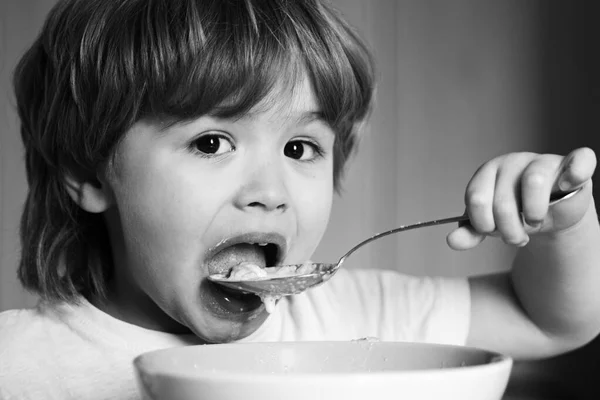 En hungrig liten pojke som äter. Glada barn äter mat själv med sked. Trevlig barnfrukost. Barnet äter mat på köket. Lycklig pojke äter hälsosam mat sked själv. — Stockfoto