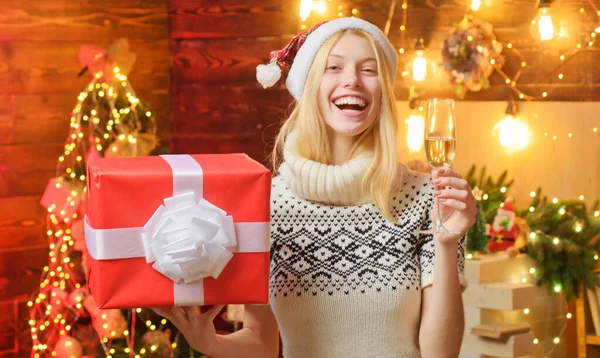 Gelukkige blonde vrouw in Santas rode hoed met een geschenk in een hande en een weduwe champagne in een ander in prachtig versierd interieur. Viering van nieuwjaarsconcept. — Stockfoto