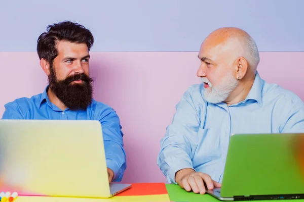 Old and young men looking at laptop screen watching movie or news in social network. Two business people. Business people having meeting around table in modern color office.