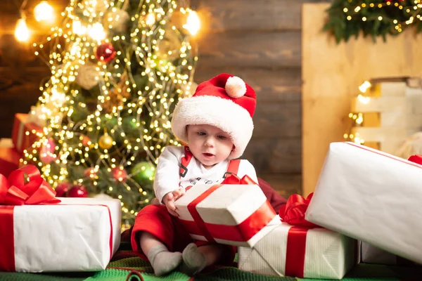 Nettes Babygesicht mit Weihnachtsmütze in der Nähe des Weihnachtsbaums. Happy Childhood, Kind 1 Jahr alt halten Geschenk. Neujahrskinder. — Stockfoto