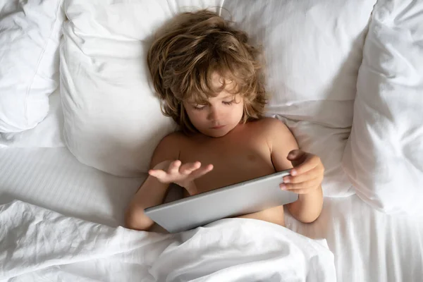 Kind mit Tablette im Bett. Kleiner Junge auf dem Sofa liegend, Film auf Tablet, im Wohnzimmer, Erlaubnis der Eltern, sicheres Online-Internet für das Kind. Junge beim Spielen. Soziales und technologisches Konzept. — Stockfoto