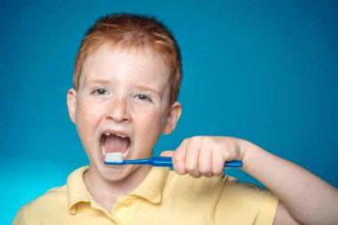 Çocuk dişlerini fırçalıyor. Dişlerini fırçalayan mutlu çocuk. Mavi arka planda diş fırçası olan tek dişli gülen çocuk. Gömlek tasarımı, sağlık, ağız temizliği..