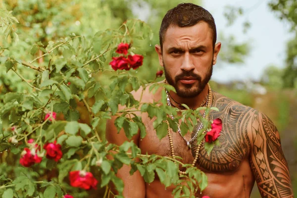 Sensual men. Handsome sexy topless male model with beautiful eyes. Sexy masculine man shirtless outdoors against roses. Sensual portrait of a very muscular male model in sensual pose.