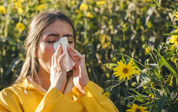 Cold flu season, runny nose. Flowering trees in background. Young girl sneezing and holding paper tissue in one hand and flower bouquet in other. Flu. Allergy season.