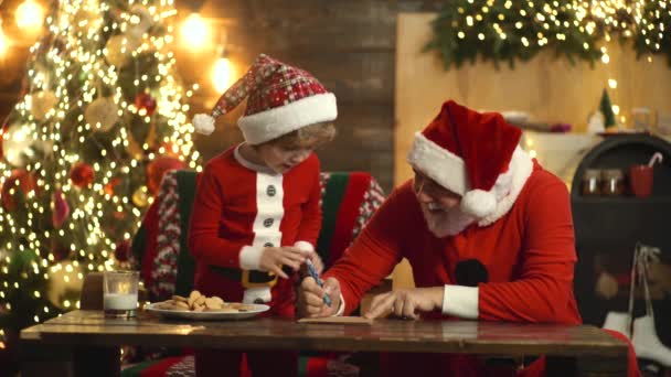 O avô e o neto celebram o Natal e o Ano Novo no chapéu de Santa. Ajudante de Natal e Santa leitura lista de desejos para casa perto da árvore de Natal. — Vídeo de Stock