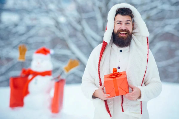 圣诞贺卡。雪园里英俊的嬉皮士带着礼物的冬季肖像使雪人成为雪人.在冬季公园里和雪人一起玩红色礼物的人. — 图库照片