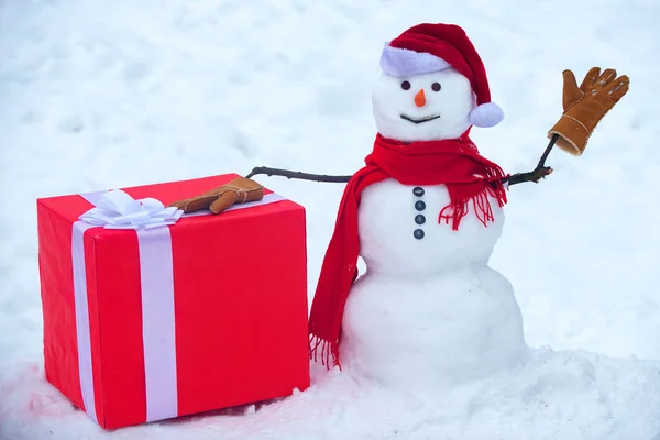 Boneco de neve o amigo está de pé em chapéu de inverno e cachecol com nariz vermelho. Boneco de neve de Natal em fundo de neve branco. Homem de neve com presente. — Fotografia de Stock