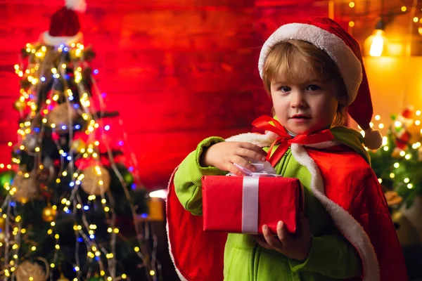 Cute child opening a Christmas present. Cheerful little boy dressed as Santa Claus. A boy in Santa hat helps with Christmas gift in a red box. Stock Image