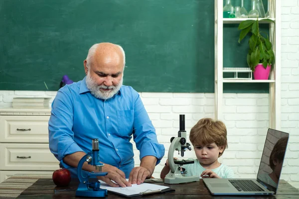 Niño amigable con viejo maestro maduro en el aula cerca del escritorio de pizarra. Retrato del abuelo y nieto en pizarra en el aula. — Foto de Stock