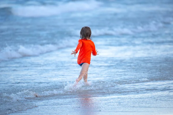 ブロンドの男の子が走っていて、夏に青い海のビーチで楽しんでいます. — ストック写真