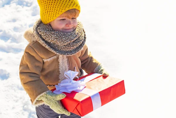 冬服の子供はクリスマスプレゼントを持っています。雪の多い冬の公園を散歩中の子供。美しい冬の自然。素晴らしい冬の公園。かわいい顔をした愛らしい子供。空のコピースペースでの写真. — ストック写真