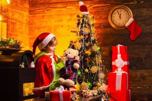 Niño en traje de Santa sosteniendo oso de peluche y esperando la medianoche de Año Nuevo. Hermoso árbol decorado de Navidad. Regalos y regalos de vacaciones de invierno. Concepto de infancia feliz. — Foto de Stock