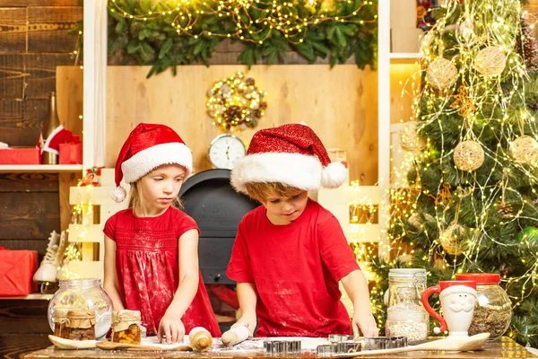 Crianças bonitas em chapéus de Papai Noel preparação jantar de férias para a família. Dois rapazes giros com chapéu de Pai Natal. Duas crianças em chapéus e aventais de Papai Noel preparando biscoitos de Natal juntos. — Fotografia de Stock