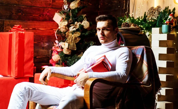 Mutlu Noeller ve mutlu yıllar. Yakışıklı Noel Baba. Moda adamı elinde bir hediyeyle Noel ağacının yanında oturuyor. Kış, tatil konsepti. — Stok fotoğraf