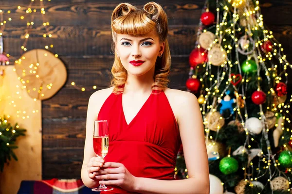 Portret van een mooie vrolijke vrouw in rode kerstjurk met champagneglas terwijl ze staat en omhoog kijkt over de achtergrond van de kerstboom. — Stockfoto