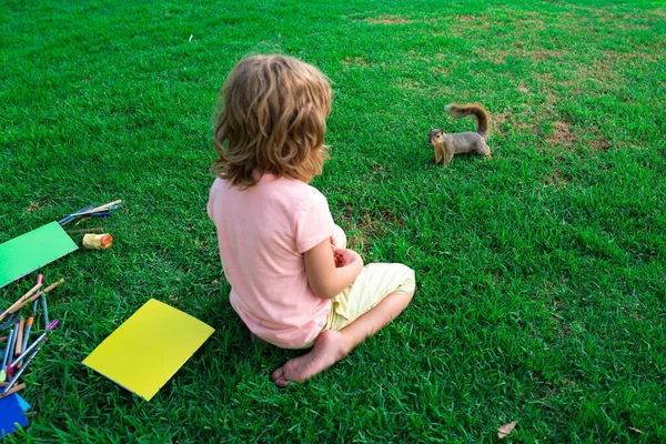 Kind leerling zittend op het gazon in het park, spelen met eekhoorn. Studeren en buiten leren. Kind bereidt zich voor op school buiten. — Stockfoto