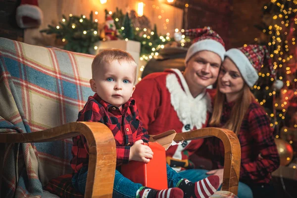 待望の子供。子供はロッキングチェアに座ってカメラを見ている。幸せな家族だ。クリスマスツリーと新年の装飾. — ストック写真