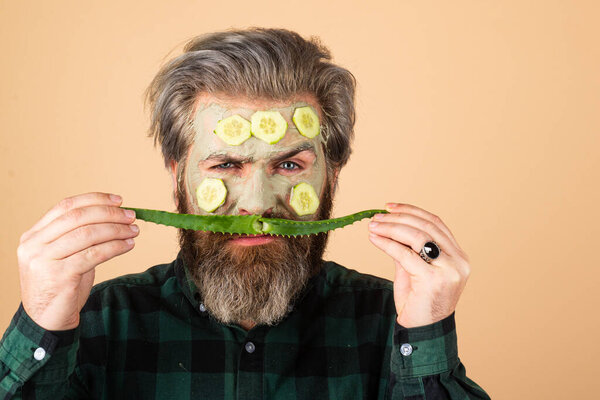 Facial treatment. Funny bearded man with clay facial mask in beauty spa. Fun guy with facial mask with Cucumber and aloe vera.