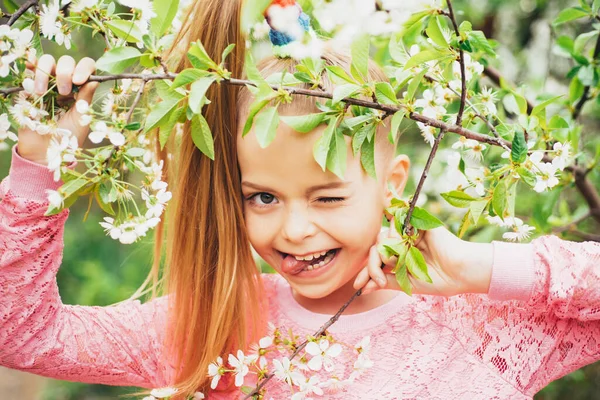 Close Up portret van klein meisje knipoogt in roze shirt met bloemen. Hij liet haar tong zien. Voorjaarsmagie. Tuinboom bloemen. Vrije tijd. — Stockfoto