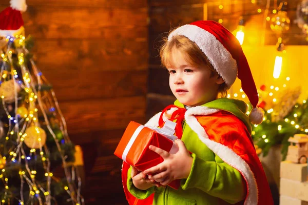 Glad liten pojke vid granen med sin julklapp. Grabben har tomtkläder på sig. Julkoncept. — Stockfoto