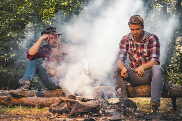 Ομάδα φίλων απολαμβάνουν πικνίκ στο δάσος και πίνοντας μπύρα. Ομάδα από δύο άντρες φίλους που κατασκηνώνουν με ζαχαρωτά πάνω από μια φωτιά σε στρατόπεδο. Ο άνθρωπος κάνει τη δουλειά του. Δύο φίλοι τύπος κάμπινγκ στο δάσος. — Φωτογραφία Αρχείου
