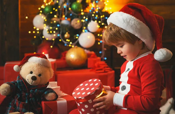 Счастливый маленький улыбающийся мальчик с рождественской подарочной коробкой. Концепция праздников. Малыш в одежде Санты. Мишка Тедди и елка. — стоковое фото