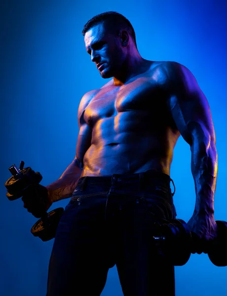 Musclé bodybuilder gars faire des exercices avec haltères isolés sur néon bleu. Athlète nu avec un corps fort. Chausson torse nu avec abdos parfaits. — Photo