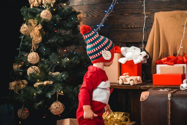 子供達よ。プレゼント付きのサンタの帽子の幸せな小さな子供たちにはクリスマスがあります。木製の背景に贈り物を持つ肖像画の子供。冬の子供たち。クリスマスツリーによって装飾的なおもちゃのボールを見て幸せな子供の肖像画. — ストック写真