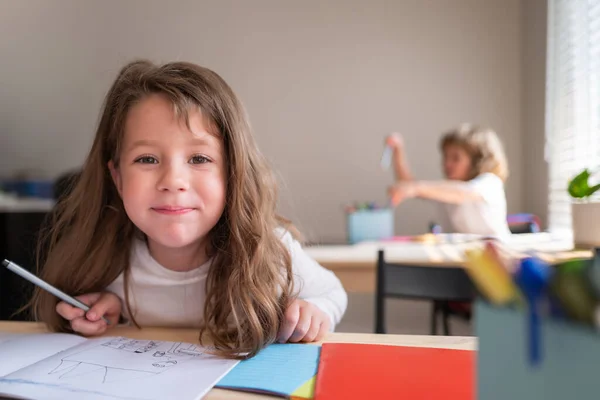 Vicces, boldog iskoláslány, aki jegyzetel az osztályban. Gyerekek a suliban az osztályteremben. Pupil az asztalnál az általános iskolában, aki kamerába néz. — Stock Fotó