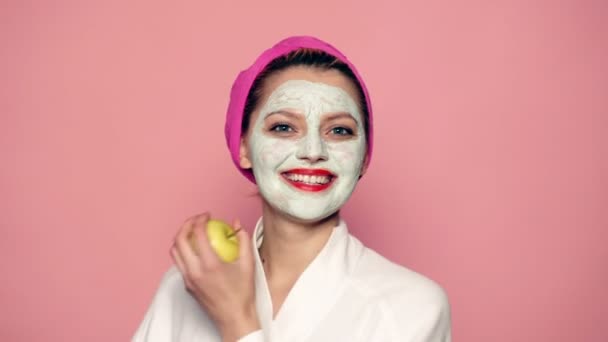 Chica con una máscara en la cara y una toalla en la cabeza sostiene una manzana en sus manos y sonríe en el fondo rosa. Concepto de cuidado de la piel. — Vídeos de Stock