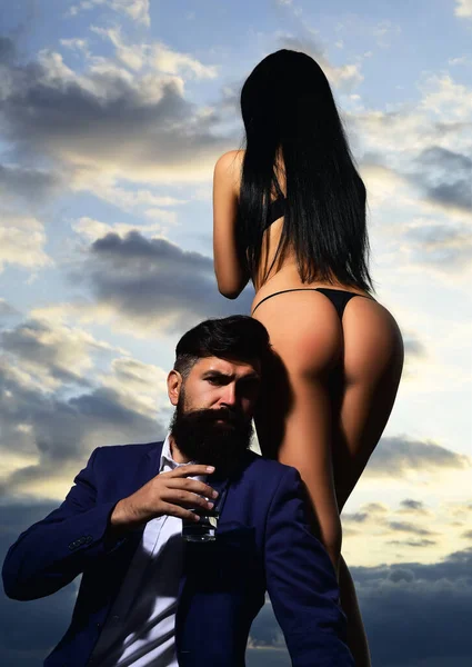 Bearded businessman in elegant suit with glass of whiskey. Rich man drink. Sencual couple in love. Womans butt in bikini. — Stock fotografie