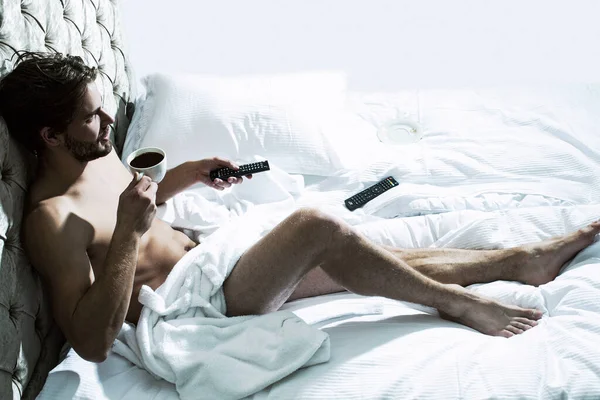 Full length gorgeous sexy nude male model laying in bed. Attractive man in bedroom drink coffee, hold remote control tv. — Stock Photo, Image