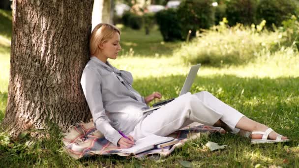 Μαθήτρια κοπέλα υπαίθρια, καυκάσια γυναίκα κάθεται στο πάρκο στο πράσινο γρασίδι με σημειωματάριο και smartphone, τα χέρια στο πληκτρολόγιο. — Αρχείο Βίντεο