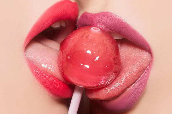 A boca feminina sensual lambe. Prazer oral lésbico. Vista de perto de dois lábios sensuais sensuais femininos brilhantes com batom rosa e roxo segurando na boca e lambendo deliciosos doces pirulito vermelho redondo. — Fotografia de Stock