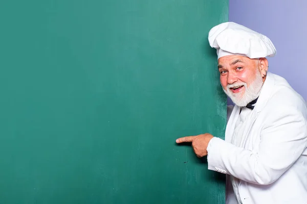 Senior chief cook. Portrait of chief cook with hand sign on blackboard background. Excited man chef cook wearing uniform showing copy space text.
