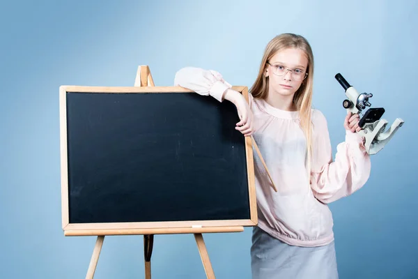 Tonårstjej i skolan. En attraktiv liten flicka med mikroskop. Porträtt av modern tonåring skolflicka nära svarta tavlan. — Stockfoto